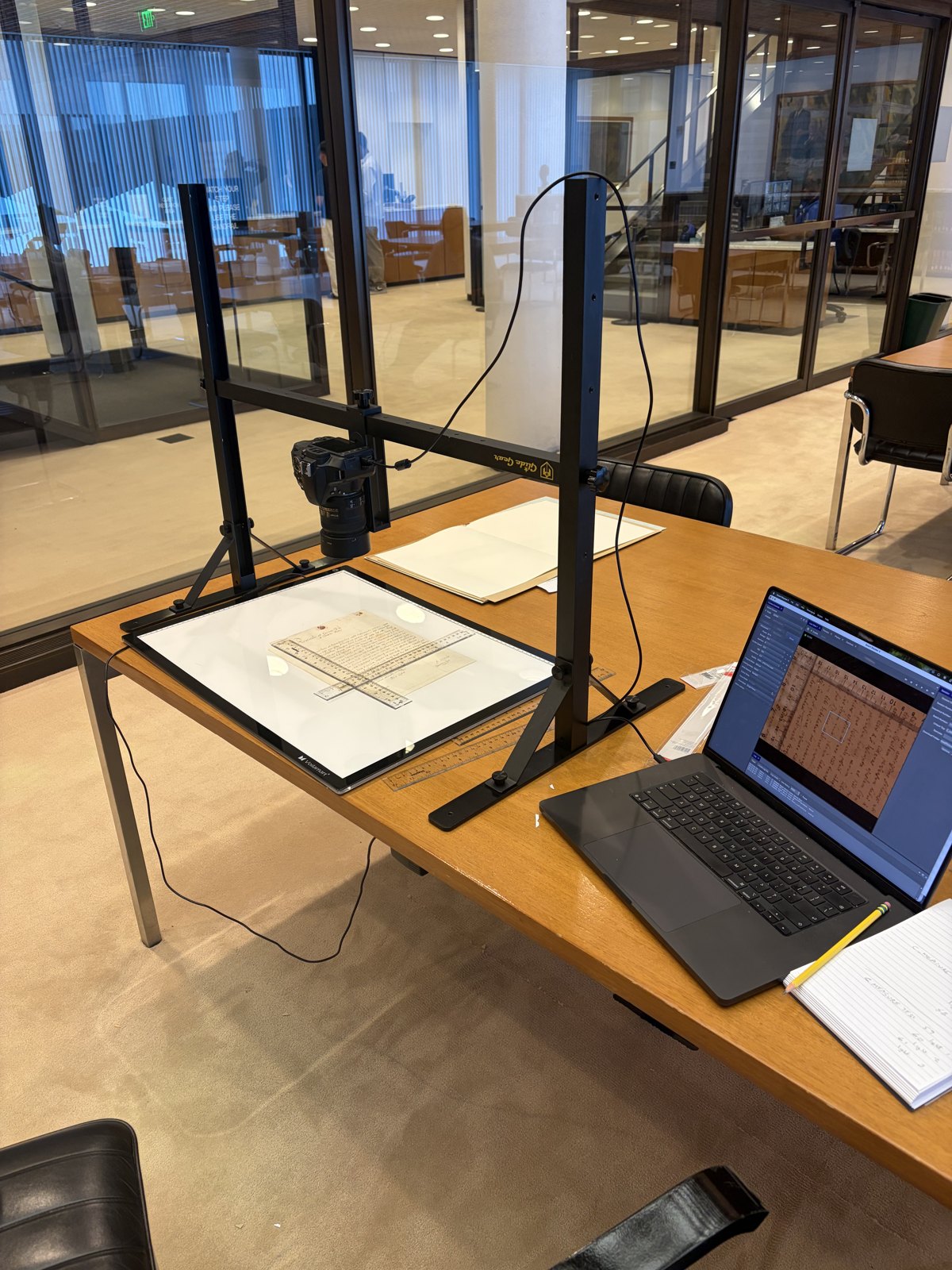 The copy stand set up on a reading-room table at the Beinecke Library: camera on the crossbar above a lit light table holding the letter and two scales, with the tethering laptop alongside.